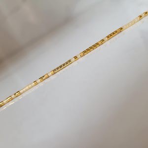 Op de afbeelding: Een delicate ketting met een streng kleine, cr&egrave;mekleurige kralen afgewisseld met goudkleurige vierkante kralen. De kralen zijn aan elkaar geregen, waardoor een lineair ontwerp ontstaat. De ketting wordt gepresenteerd tegen een witte achtergrond.