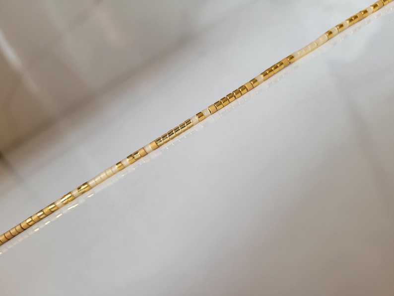 Op de afbeelding: Een delicate ketting met een streng kleine, cr&egrave;mekleurige kralen afgewisseld met goudkleurige vierkante kralen. De kralen zijn aan elkaar geregen, waardoor een lineair ontwerp ontstaat. De ketting wordt gepresenteerd tegen een witte achtergrond.