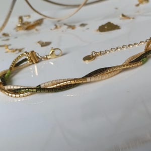May include: Gold-toned bracelet with three intertwined strands. The strands feature small, rectangular beads in gold, cream, and olive green. The bracelet has a lobster clasp closure and an adjustable chain.