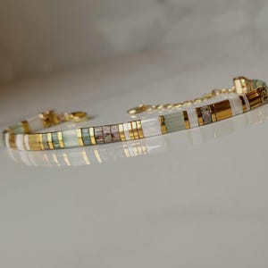 Może przedstawiać: Delikatna bransoletka ze złotym łańcuszkiem i serią prostokątnych koralików. Koraliki naprzemiennie białe, złote i jasnozielone, z niektórymi sekcjami prezentującymi wzór mozaiki. Bransoletka została zaprojektowana z myślą o wygodnym dopasowaniu.