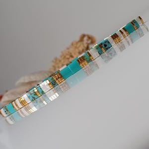 Op de afbeelding: Een turquoise, witte en gouden kralenarmband. De armband bestaat uit kleine, rechthoekige kralen in afwisselende kleuren. De gouden kralen hebben een metallic glans. De armband wordt gepresenteerd op een reflecterend oppervlak.