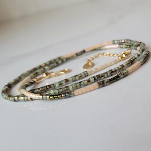 May include: A delicate necklace with small, square beads in green, beige, and gold. The necklace has a gold-coloured clasp and chain, and is likely made of glass or a similar material. The beads are strung together to create a layered effect.