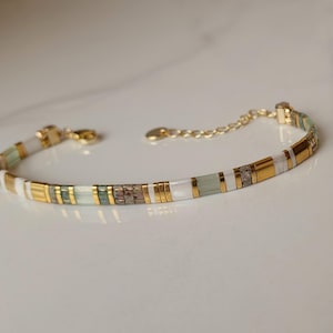 Puede incluir: Una pulsera delicada con un patr&oacute;n repetitivo de cuentas cil&iacute;ndricas en oro met&aacute;lico, blanco opaco, verde salvia p&aacute;lido y bronce texturizado. La pieza incluye un cierre de mosquet&oacute;n en tono dorado y una cadena ajustable con una peque&ntilde;a etiqueta.