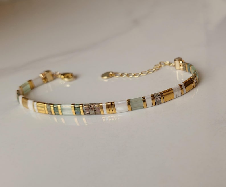 Puede incluir: Una pulsera delicada con un patr&oacute;n repetitivo de cuentas cil&iacute;ndricas en oro met&aacute;lico, blanco opaco, verde salvia p&aacute;lido y bronce texturizado. La pieza incluye un cierre de mosquet&oacute;n en tono dorado y una cadena ajustable con una peque&ntilde;a etiqueta.