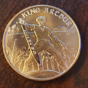 May include: A gold-colored coin with a raised image of King Arthur holding a sword. The coin has the words "KING ARTHUR" inscribed around the edge.