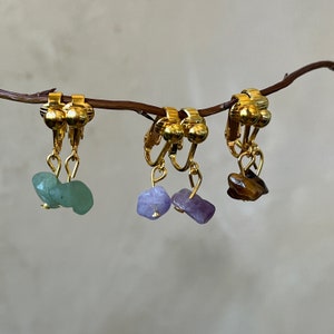 Puede incluir: Tres pares de pendientes de clip dorados con acentos de piedras preciosas de diferentes colores. Los pendientes presentan una piedra verde, morada y marrón.