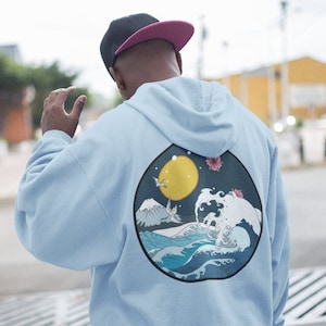 Op de afbeelding: Een lichtblauwe hoodie met een grafische print van een golf, een berg, een maan en een vogel op de rug. De grafiek bevindt zich in een cirkel met een donkerblauwe omtrek.