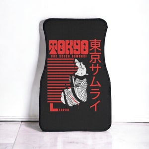 May include: Black car floor mat featuring a red and white graphic of a geisha and the text "TOKYO THE SEVEN SAMURAI" in red. Japanese text is also present. The mat has a rectangular shape with rounded edges.