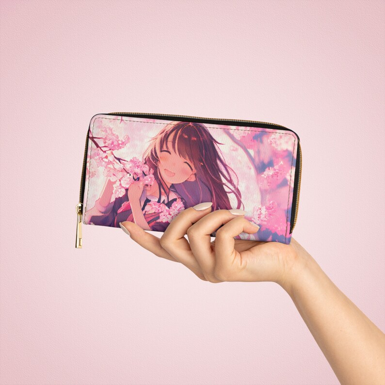 Puede incluir: Una cartera con cremallera rosa y blanca con una ilustraci&oacute;n de anime colorida de una chica con el pelo largo y casta&ntilde;o sonriendo y sosteniendo flores rosas. El fondo es un patr&oacute;n floral rosa y blanco.