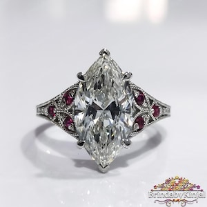 May include: A marquise-cut diamond engagement ring with a detailed filigree band. The ring features a large, clear center stone and is accented with small red gemstones. The ring is set on a white surface.