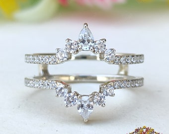 May include: A silver ring with a pear-shaped diamond in the center and smaller diamonds surrounding it. The ring is designed to be worn as a wedding band enhancer.