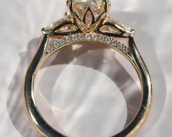 Puede incluir: Un anillo de compromiso dorado con una piedra central grande y transparente y piedras laterales más pequeñas. La banda pulida tiene una fila de pequeños diamantes. El anillo está sobre un fondo blanco suave.