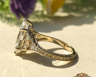 Op de afbeelding: Een gouden verlovingsring met een grote, heldere, peervormige edelsteen. De ring heeft ingewikkelde filigraan en kleine accentstenen op de band en de zetting. De ring staat tegen een zachte achtergrond met bloemen.
