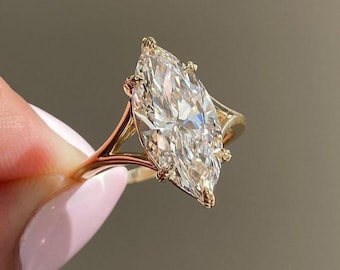 Könnte beinhalten: Ein Verlobungsring mit Diamanten im Marquiseschliff und einem goldenen Band. Der große, klare Diamant ist der Mittelpunkt, der mit zarten Zinken gefasst ist. Das goldene Band hat ein geteiltes Schaftdesign, das zur Eleganz des Rings beiträgt. Der Ring wird hochgehalten, um sein Funkeln zu zeigen.