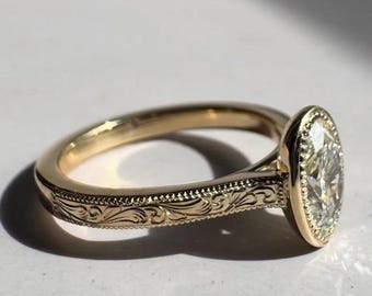 May include: A gold ring with an oval-shaped diamond. The ring has an engraved floral design on the band and a bezel setting. The diamond is clear and sparkling, and the ring is set against a white background.