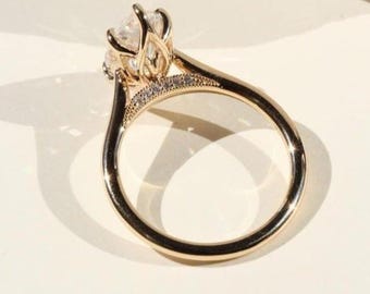 May include: A gold engagement ring with a large, clear gemstone at the center. The ring features a delicate band with small accent stones and a detailed setting. The ring is displayed on a white surface, showcasing its intricate design.