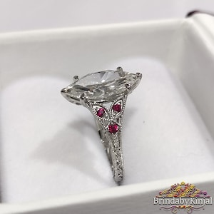 May include: A marquise-cut diamond ring with a detailed silver band. The ring features three small red gemstones and intricate filigree designs. The ring is set in a white box.