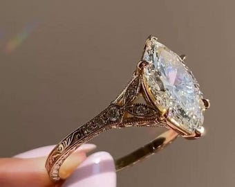 以下が含まれることがあります： 大きなペアシェイプのダイヤモンドが付いたゴールドの婚約指輪。バンドには複雑なフィリグリーと小さなアクセントダイヤモンドがあります。リングは柔らかくニュートラルな背景に対して保持されています。