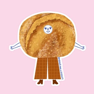 May include: A whimsical sticker featuring a smiling, anthropomorphic cinnamon roll character. The character has a round, textured body, arms, and is wearing brown checkered pants and shoes. The text "LINDSEY DAY STUDIO" is printed vertically on the right side.