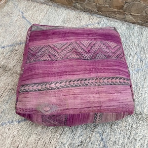 May include: A square floor pouf featuring a vibrant magenta and purple woven textile with horizontal bands of intricate geometric patterns, including zigzags and diamonds, crafted from a textured fabric with visible stitching along the edges.