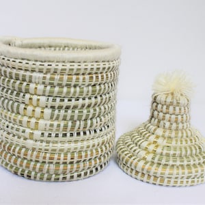 May include: A woven storage basket with a lid. The basket is made of natural materials and has a white and gold striped pattern. The lid has a white pom-pom on top.