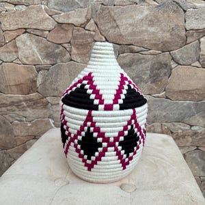 Puede incluir: Una cesta tejida blanca con un patrón geométrico rosa y negro. La cesta tiene una base redonda y una parte superior ligeramente puntiaguda.