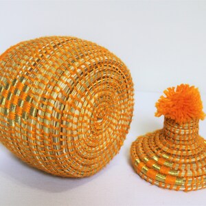 Peut inclure: Un panier rond en osier avec un couvercle. Le panier est fait de fibres naturelles et pr&eacute;sente une combinaison de couleurs orange vif et or. Le couvercle est surmont&eacute; d'un pompon orange moelleux.