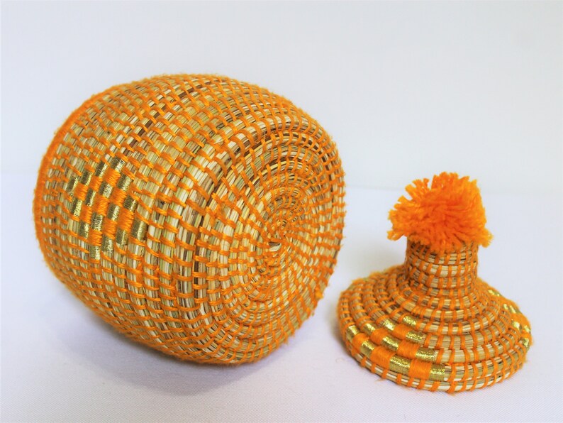 Peut inclure: Un panier rond en osier avec un couvercle. Le panier est fait de fibres naturelles et pr&eacute;sente une combinaison de couleurs orange vif et or. Le couvercle est surmont&eacute; d'un pompon orange moelleux.