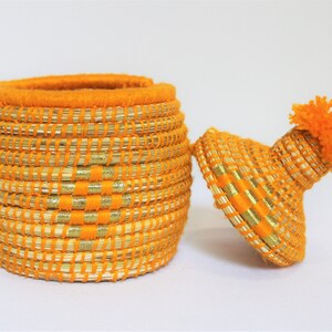 Peut inclure: Un panier de rangement tiss&eacute; orange et or avec un couvercle et un pompon sur le dessus.