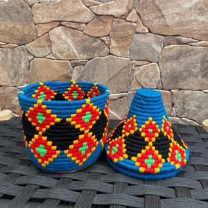 May include: A cylindrical container with a matching conical lid, both crafted from blue, red, yellow, and green yarn. The exterior features a repeating geometric diamond pattern in black, red, and yellow, with small green crosses at the center.