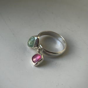 Op de afbeelding: Een zilveren ring met twee edelsteenhangers. Eén hanger is lichtgroen, de andere roze. De ring staat tegen een witte achtergrond.