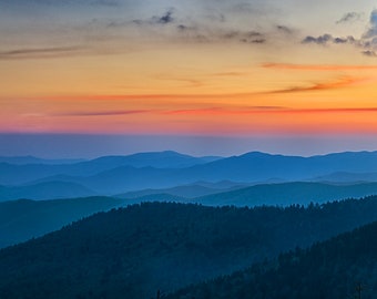 May include: A panoramic view of a mountain range at sunset. The sky is a vibrant orange and pink, with wispy clouds. The mountains are a deep blue, with a hazy, layered effect.