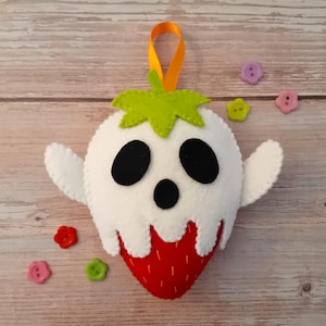 May include: A felt strawberry ghost ornament featuring a red strawberry base with white felt dripping over the top, black felt eyes and mouth, a green felt stem, and an orange ribbon loop for hanging, accompanied by several small colourful buttons.