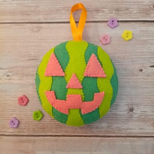 May include: A round felt ornament shaped like a watermelon with a jack-o'-lantern face. The body features alternating light and dark green stripes with pink triangular eyes, a small pink nose, and a wide pink mouth. A yellow ribbon loop is attached.