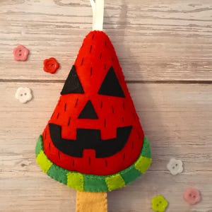 May include: A felt Halloween decoration in the shape of a watermelon slice with a carved jack-o'-lantern face. The red felt has black triangle eyes and a carved mouth. It has a green and yellow striped rind and a tan felt stick. A white ribbon is attached for hanging.