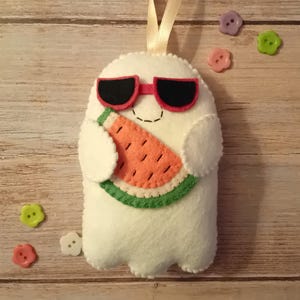 May include: A white felt ghost ornament with a cream ribbon loop for hanging. The ghost wears red-framed black sunglasses and holds a felt watermelon slice with an orange centre, black seeds, and a green rind. Small colourful buttons surround it.