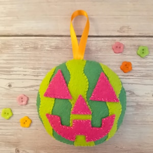 May include: A felt pumpkin decoration with a green and yellow striped design. The face is made of pink felt and has a yellow ribbon for hanging. Small decorative buttons in pink, orange, and green are scattered around the pumpkin.