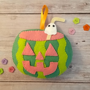 May include: A felt watermelon jack-o'-lantern ornament featuring a green rind, pink triangular eyes, a pink triangular nose, and a pink jagged mouth. A white ghost with black eyes sits on top with a straw, and a yellow ribbon loop is attached.