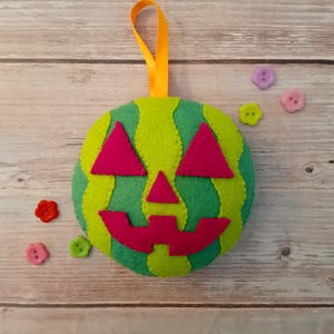 May include: A round, felt pumpkin ornament featuring alternating vertical stripes of light green and dark green. It has a jack-o'-lantern face made of dark pink felt, including two triangular eyes, a triangular nose, and a jagged mouth, with an orange ribbon loop.