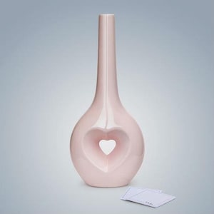 Può includere: Un vaso in ceramica rosa chiaro con un'apertura a forma di cuore. Il vaso ha un collo stretto e una base larga.