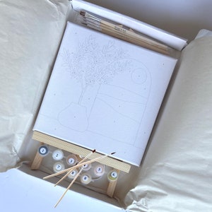 Pode incluir: Um kit de pintura por n&uacute;meros com uma tela branca e um desenho em preto e branco de uma oliveira num vaso. O kit inclui nove potes de tinta, tr&ecirc;s pinc&eacute;is e um cavalete de madeira.