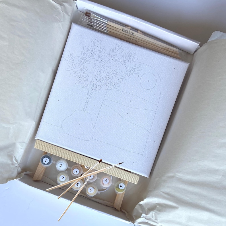 Pode incluir: Um kit de pintura por n&uacute;meros com uma tela branca e um desenho em preto e branco de uma oliveira num vaso. O kit inclui nove potes de tinta, tr&ecirc;s pinc&eacute;is e um cavalete de madeira.