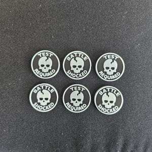 May include: Six circular patches arranged in two rows of three. Each patch is black with a white skull graphic featuring a crack on the forehead. Three patches display the text TEST REQUIRED, and three patches display the text BATTLE SHOCKED.