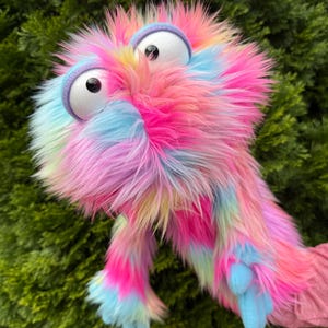 May include: A hand puppet covered in long, shaggy faux fur featuring a vibrant, multi-colored pattern of bright pink, light blue, and yellow. The puppet has two large, round white eyes with black pupils, framed by light purple fabric eyelids.