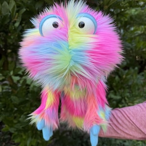 May include: A hand puppet covered in long, shaggy faux fur featuring a vibrant tie-dye pattern of pink, yellow, and light blue. The puppet has two large, round white eyes with black pupils, framed by light blue circular rims, and two light blue hands.