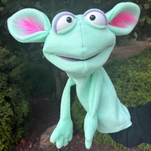 May include: A mint green hand puppet featuring large, rounded ears with bright pink faux fur accents. The puppet has white eyes with black pupils, purple eyelids, and a wide, purple-lined mouth. It includes two long arms with three-fingered hands.