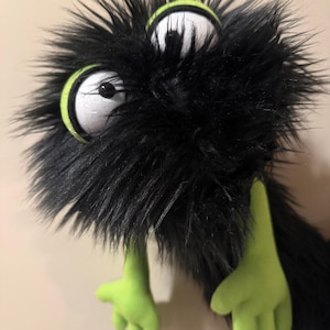 May include: A plush monster with black fur, large white eyes with black pupils, and lime green accents. The monster has two lime green hands and a fluffy, textured appearance. The eyes are outlined in lime green.