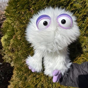 May include: A fluffy white puppet with large, round eyes and purple accents. The puppet has a shaggy texture and small, purple feet. The puppet is held in front of a green bush.