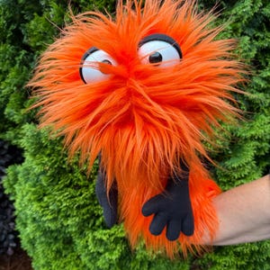 May include: Orange, furry puppet with large white eyes, black pupils, and a black outline. The puppet has black hands and is set against a backdrop of green foliage. The puppet's fur is long and shaggy.