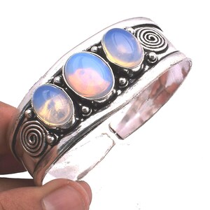 Könnte beinhalten: Ein silberfarbenes Armband mit drei ovalen, durchscheinenden, irisierenden Cabochon-Steinen in einer Reihe. Das Metallband ist an beiden Enden mit geprägten Spiralmustern und kleinen kugelförmigen Akzenten zwischen den Steinen verziert.